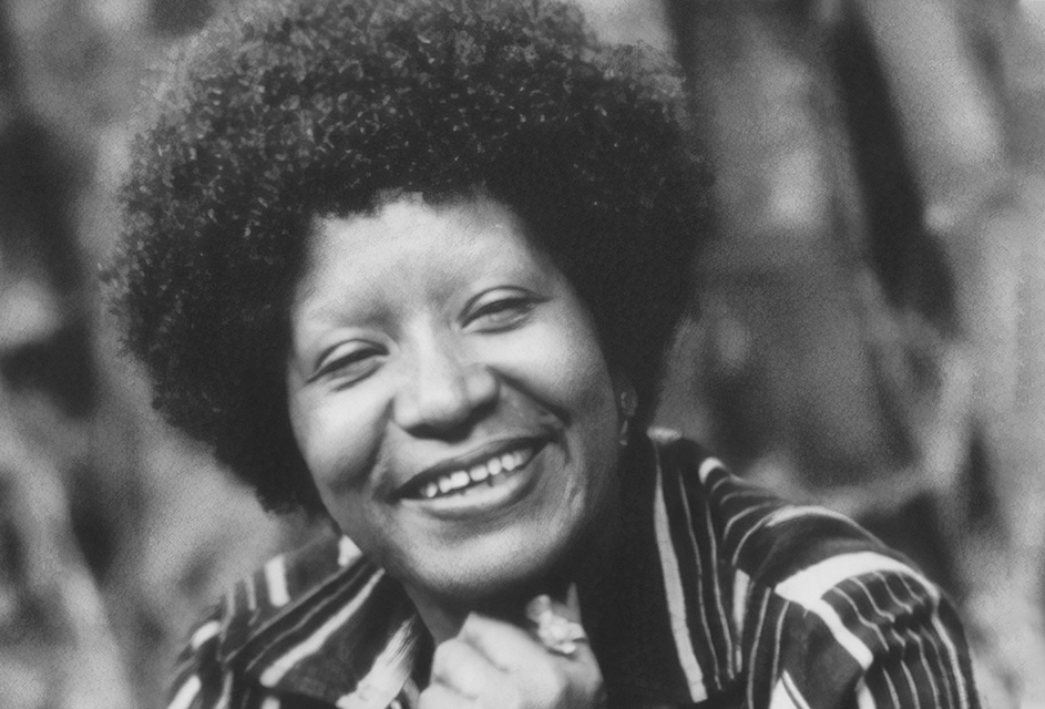 Lélia Gonzalez Sorrindo em um retrato preto e branco, é uma mulher negra com cabelos crespos longos